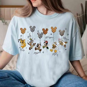 May include: Light blue t-shirt featuring Disney characters Pluto, Goofy, Mickey Mouse, Minnie Mouse, Donald Duck, and Daisy Duck. Each character is holding a Mickey Mouse-shaped balloon with a unique pattern. Signatures are below each character.