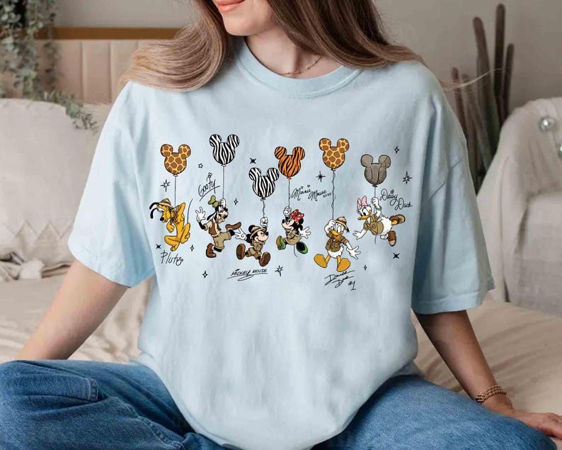 May include: Light blue t-shirt featuring Disney characters Pluto, Goofy, Mickey Mouse, Minnie Mouse, Donald Duck, and Daisy Duck. Each character is holding a Mickey Mouse-shaped balloon with a unique pattern. Signatures are below each character.