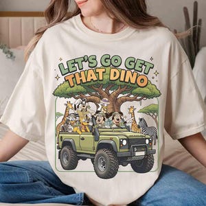 May include: Cream-colored t-shirt featuring a cartoon safari scene with the text "LET'S GO GET THAT DINO." The design includes a green jeep filled with cartoon characters, a tree, giraffes, and a zebra.