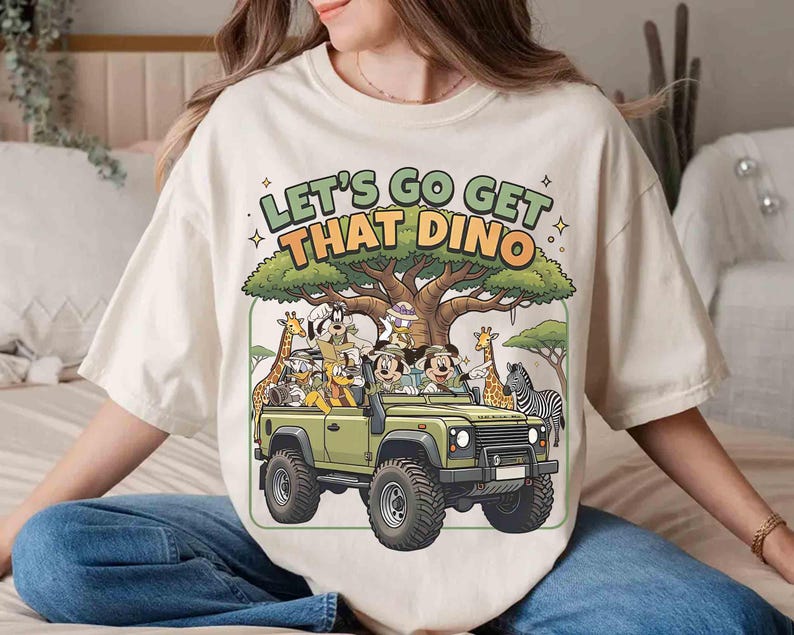 May include: Cream-colored t-shirt featuring a cartoon safari scene with the text "LET'S GO GET THAT DINO." The design includes a green jeep filled with cartoon characters, a tree, giraffes, and a zebra.