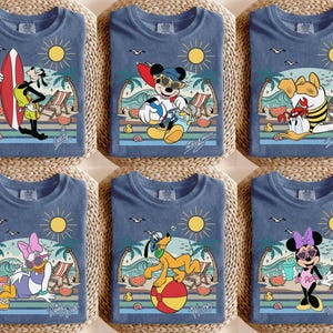 K&ouml;nnte beinhalten: Sechs blaue T-Shirts mit farbenfrohen Strandmotiven mit Disney-Figuren. Jedes Shirt hat eine andere Figur: Goofy mit einem Surfbrett, Mickey Mouse mit einem Beachball, Donald Duck, Daisy Duck, Pluto und Minnie Mouse. Die Grafiken zeigen Sonne, Wellen und Palmen.