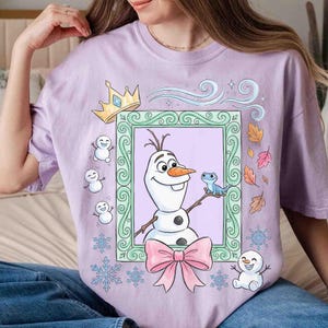 May include: A light purple t-shirt featuring a graphic print of Olaf the snowman inside a light green ornate frame. The design includes a yellow crown, a small blue lizard, three tiny snowmen, falling autumn leaves, and blue snowflakes.