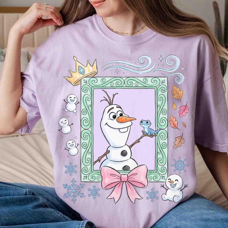 May include: A light purple t-shirt featuring a graphic print of Olaf the snowman inside a light green ornate frame. The design includes a yellow crown, a small blue lizard, three tiny snowmen, falling autumn leaves, and blue snowflakes.