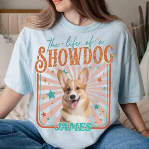 May include: Light blue t-shirt featuring a smiling Corgi dog graphic with the text "the life of a SHOWDOG" and the name "JAMES". The design includes a retro-style sunburst and star accents.