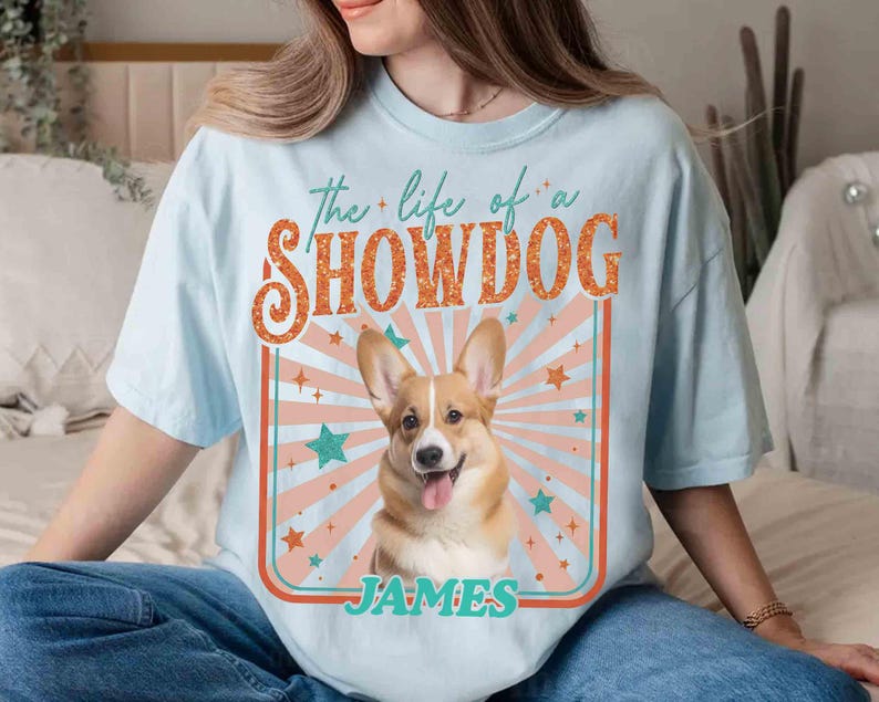 May include: Light blue t-shirt featuring a smiling Corgi dog graphic with the text "the life of a SHOWDOG" and the name "JAMES". The design includes a retro-style sunburst and star accents.