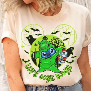 May include: Cream-colored t-shirt featuring a green cartoon character resembling Stitch, dressed as Oogie Boogie, holding a rag doll. The design includes bats, ghosts, and the text "Let's Oogie Boogie" in green, set against a Halloween-themed background.