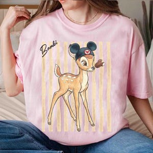 May include: A light pink t-shirt featuring a graphic of the character Bambi wearing a black mouse-ear hat. The design includes vertical beige stripes in the background and the word Bambi written in black cursive script to the left of the character.