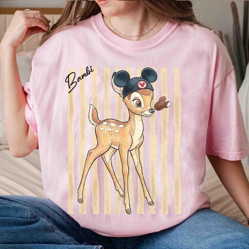 May include: A light pink t-shirt featuring a graphic of the character Bambi wearing a black mouse-ear hat. The design includes vertical beige stripes in the background and the word Bambi written in black cursive script to the left of the character.