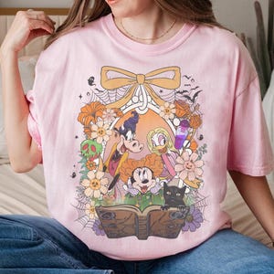 K&ouml;nnte beinhalten: Hellrosa T-Shirt mit einem farbenfrohen Aufdruck von Zeichentrickfiguren, Blumen und einem Zauberbuch. Das Design umfasst eine schwarze Katze, Flederm&auml;use und eine Schleife. Das Shirt besteht aus einem weichen Material und eignet sich f&uuml;r Freizeitkleidung.