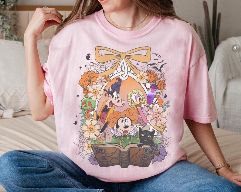 K&ouml;nnte beinhalten: Hellrosa T-Shirt mit einem farbenfrohen Aufdruck von Zeichentrickfiguren, Blumen und einem Zauberbuch. Das Design umfasst eine schwarze Katze, Flederm&auml;use und eine Schleife. Das Shirt besteht aus einem weichen Material und eignet sich f&uuml;r Freizeitkleidung.