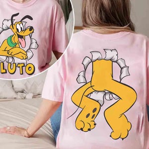 May include: Pale pink t-shirt with a cartoon Pluto design. The front shows Pluto's face emerging from a torn paper effect, with the name "PLUTO" below. The back of the shirt features Pluto's legs and paws bursting through a similar design.