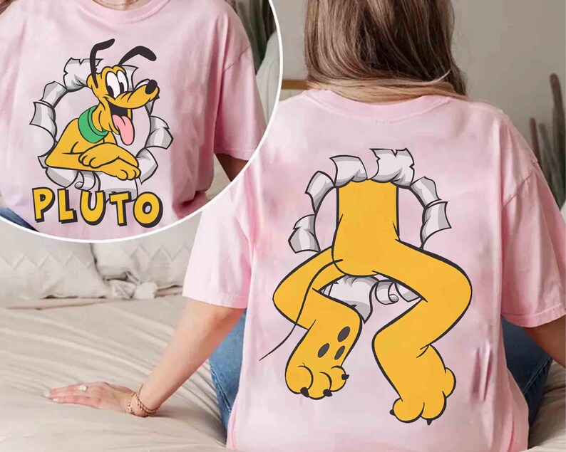 May include: Pale pink t-shirt with a cartoon Pluto design. The front shows Pluto's face emerging from a torn paper effect, with the name "PLUTO" below. The back of the shirt features Pluto's legs and paws bursting through a similar design.