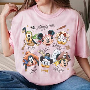 May include: Pink t-shirt featuring cartoon characters Mickey Mouse, Pluto, Donald Duck, Daisy Duck, Goofy, and Minnie Mouse in pirate attire. The shirt has the text "Mickey Mouse" and character signatures.