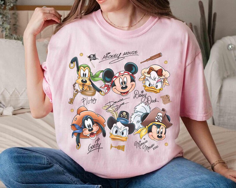 May include: Pink t-shirt featuring cartoon characters Mickey Mouse, Pluto, Donald Duck, Daisy Duck, Goofy, and Minnie Mouse in pirate attire. The shirt has the text "Mickey Mouse" and character signatures.