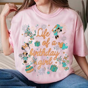 May include: A light pink t-shirt featuring a colorful graphic with the text "Life of a Birthday Girl." The design includes cartoon characters, fireworks, gifts, and a birthday cake. The shirt is a short-sleeved, crew neck style.