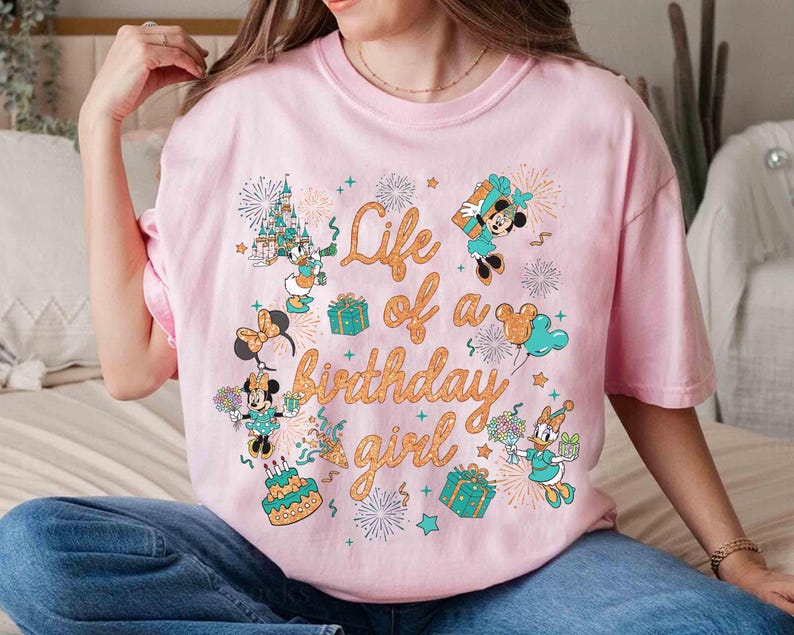 May include: A light pink t-shirt featuring a colorful graphic with the text "Life of a Birthday Girl." The design includes cartoon characters, fireworks, gifts, and a birthday cake. The shirt is a short-sleeved, crew neck style.