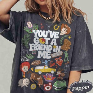 May include: Dark gray t-shirt with a colorful graphic featuring characters and text from the movie "Toy Story." The shirt includes the phrase "You've Got a Friend in Me" and various character illustrations. The shirt is labeled "Comfort Colors Pepper."