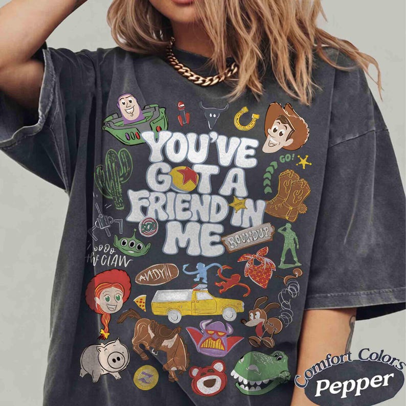 May include: Dark gray t-shirt with a colorful graphic featuring characters and text from the movie "Toy Story." The shirt includes the phrase "You've Got a Friend in Me" and various character illustrations. The shirt is labeled "Comfort Colors Pepper."