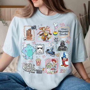 May include: Light blue t-shirt featuring a collage of Disney's Hollywood Studios characters and icons. Includes images of Sully, Darth Vader, Mickey Mouse, and the text "Disney's Hollywood Studios" and "2026".