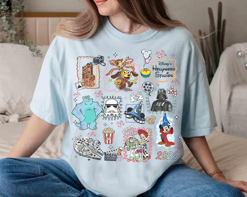 May include: Light blue t-shirt featuring a collage of Disney's Hollywood Studios characters and icons. Includes images of Sully, Darth Vader, Mickey Mouse, and the text "Disney's Hollywood Studios" and "2026".