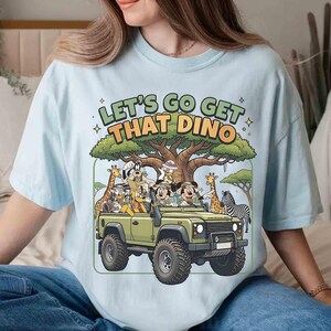 May include: Light blue t-shirt featuring a cartoon safari scene. The design includes a green jeep with cartoon characters, giraffes, and a zebra. Text on the shirt reads "LET'S GO GET THAT DINO".