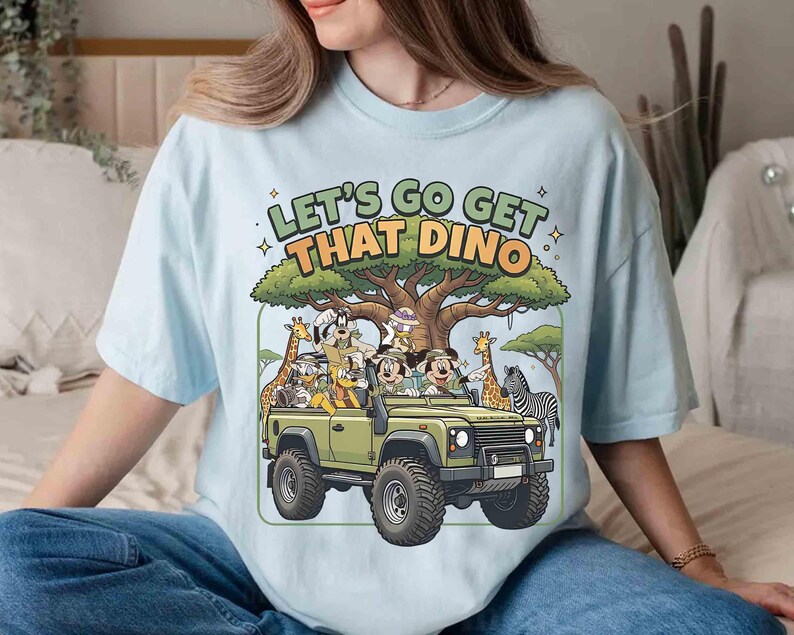 May include: Light blue t-shirt featuring a cartoon safari scene. The design includes a green jeep with cartoon characters, giraffes, and a zebra. Text on the shirt reads "LET'S GO GET THAT DINO".