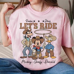May include: A light pink t-shirt featuring a rodeo theme with the text "LET'S RIDE" and illustrations of Mickey Mouse, Goofy, and Donald Duck dressed as cowboys. The shirt also includes the names "Mickey, Goofy, Donald".