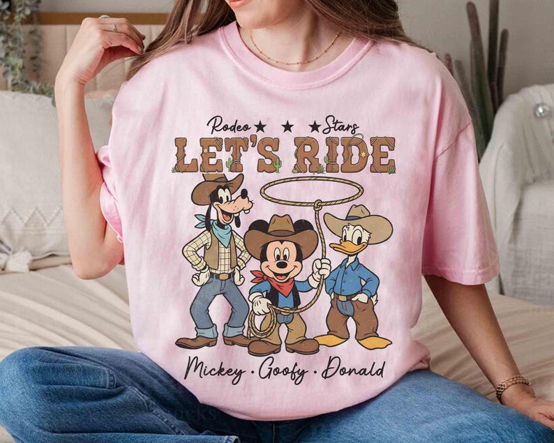 May include: A light pink t-shirt featuring a rodeo theme with the text "LET'S RIDE" and illustrations of Mickey Mouse, Goofy, and Donald Duck dressed as cowboys. The shirt also includes the names "Mickey, Goofy, Donald".
