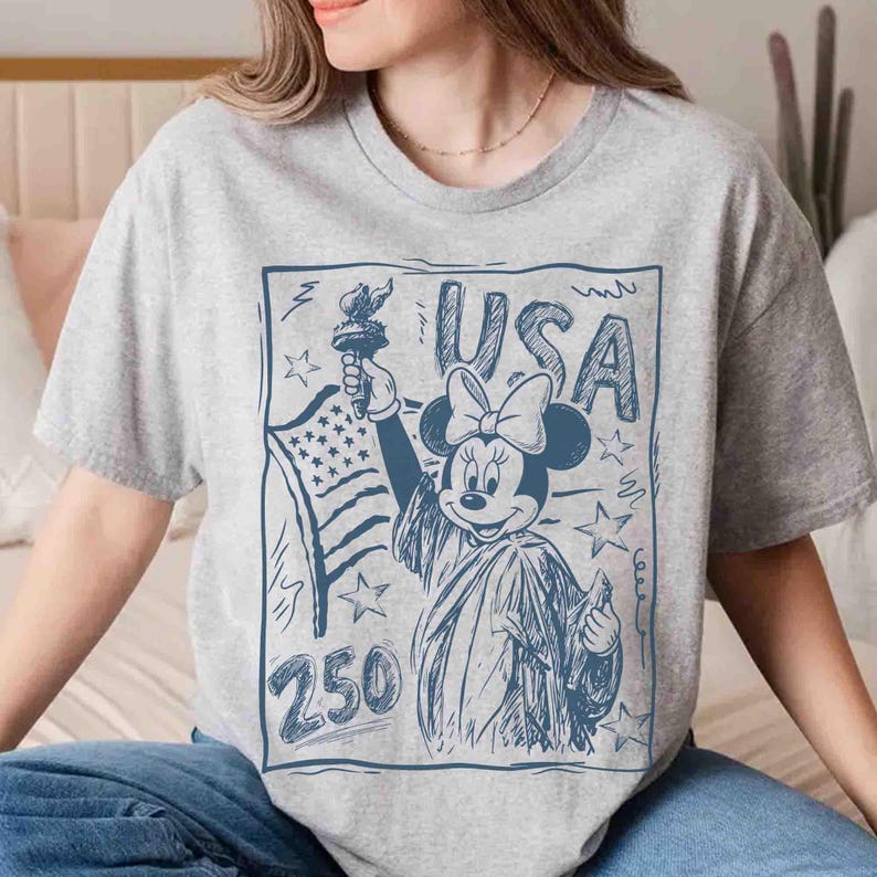 K&ouml;nnte beinhalten: Graues T-Shirt mit einer blauen Grafik von Minnie Mouse als Freiheitsstatue, die eine Fackel und die amerikanische Flagge h&auml;lt. Das Wort "USA" steht &uuml;ber Minnie, und die Zahl "250" darunter. Das Design ist im Skizzenstil gehalten.