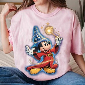 May include: A light pink t-shirt featuring a graphic of Mickey Mouse dressed as a wizard. Mickey is wearing a blue wizard hat with stars, a red robe, and holding a glowing wand. The design has a cartoon style.