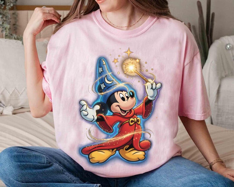 May include: A light pink t-shirt featuring a graphic of Mickey Mouse dressed as a wizard. Mickey is wearing a blue wizard hat with stars, a red robe, and holding a glowing wand. The design has a cartoon style.