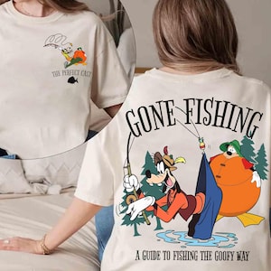 May include: Cream-colored t-shirt featuring a cartoon graphic of Goofy fishing. The front of the shirt has a small graphic with the text "THE PERFECT CAST". The back of the shirt has a larger graphic with the text "GONE FISHING" and "A GUIDE TO FISHING THE GOOFY WAY".