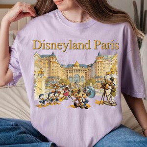 May include: A light purple t-shirt featuring the text "Disneyland Paris" in gold lettering above a colorful illustration of Disney characters in front of a building. The shirt is a comfortable, casual style.