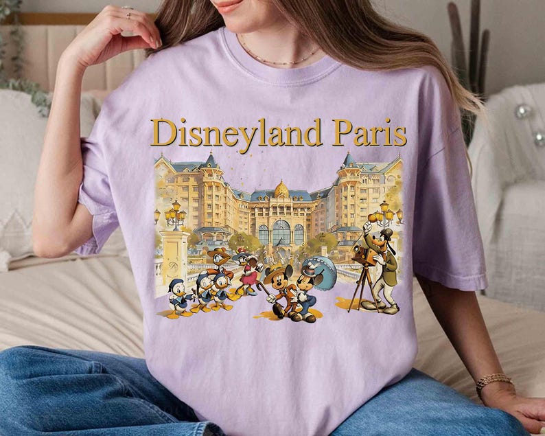 May include: A light purple t-shirt featuring the text "Disneyland Paris" in gold lettering above a colorful illustration of Disney characters in front of a building. The shirt is a comfortable, casual style.