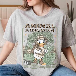 Puede incluir: Una camiseta gris con un dise&ntilde;o de Pato Donald vestido de safari, con las palabras "ANIMAL KINGDOM" en letras con estampado de leopardo. La camiseta tambi&eacute;n incluye el texto "Walt Disney World".