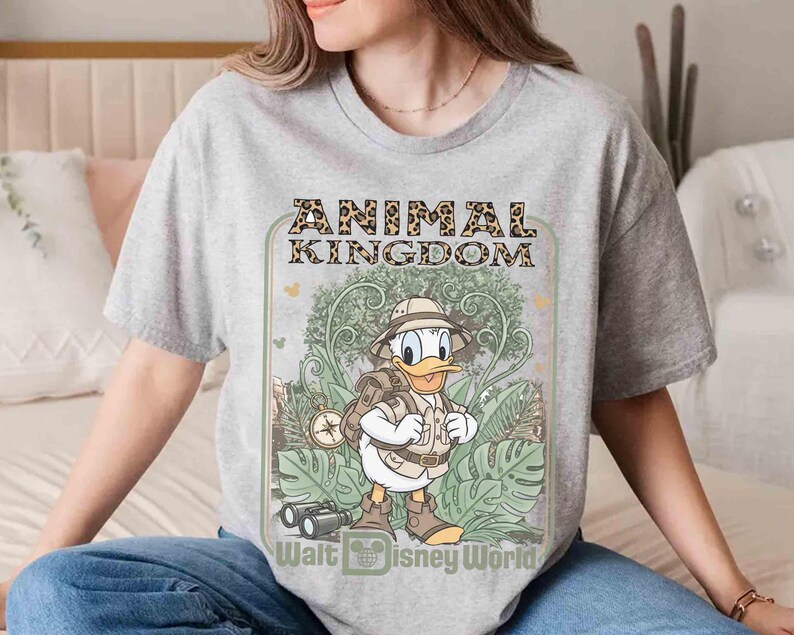Puede incluir: Una camiseta gris con un dise&ntilde;o de Pato Donald vestido de safari, con las palabras "ANIMAL KINGDOM" en letras con estampado de leopardo. La camiseta tambi&eacute;n incluye el texto "Walt Disney World".