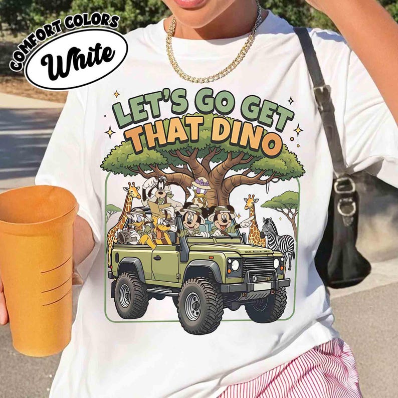 May include: White t-shirt with a colorful graphic featuring Mickey and Minnie Mouse, Donald Duck, and Goofy in a safari jeep with the text "LET'S GO GET THAT DINO". The design includes giraffes and a zebra.