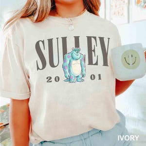 May include: Ivory-colored t-shirt featuring the word "SULLEY" above a cartoon character with blue fur and purple spots. The numbers "2001" are below the character. A white mug with a smiley face is held in the hand.