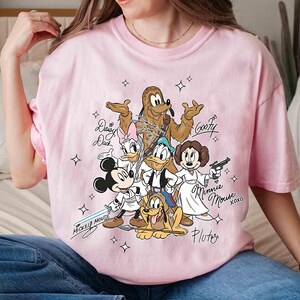 May include: Pink t-shirt featuring a graphic of Disney characters dressed as Star Wars characters. Mickey Mouse, Donald Duck, Goofy, Pluto, Daisy Duck, and Minnie Mouse are depicted with lightsabers and blasters. Text includes character names and "Mickey Mouse" and "Minnie Mouse xoxo".