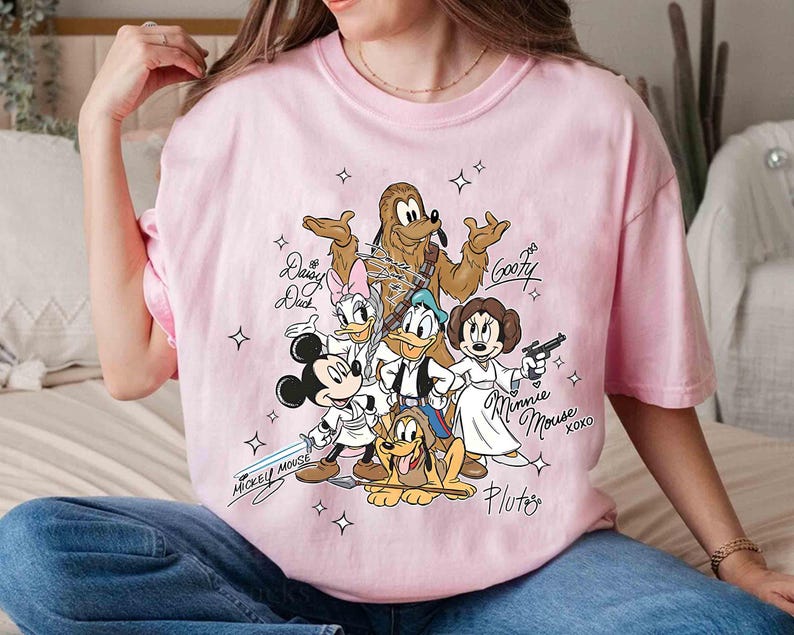 May include: Pink t-shirt featuring a graphic of Disney characters dressed as Star Wars characters. Mickey Mouse, Donald Duck, Goofy, Pluto, Daisy Duck, and Minnie Mouse are depicted with lightsabers and blasters. Text includes character names and "Mickey Mouse" and "Minnie Mouse xoxo".