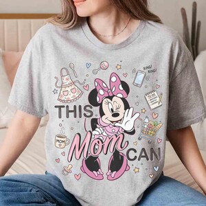 K&ouml;nnte beinhalten: Ein graues T-Shirt mit einem Minnie Mouse-Motiv und dem Text "THIS MOM CAN". Das Design umfasst eine rosa Schleife, eine Sch&uuml;rze, einen Einkaufswagen, ein Telefon und andere Symbole. Das Shirt ist ein l&auml;ssiges Kleidungsst&uuml;ck f&uuml;r den Alltag.