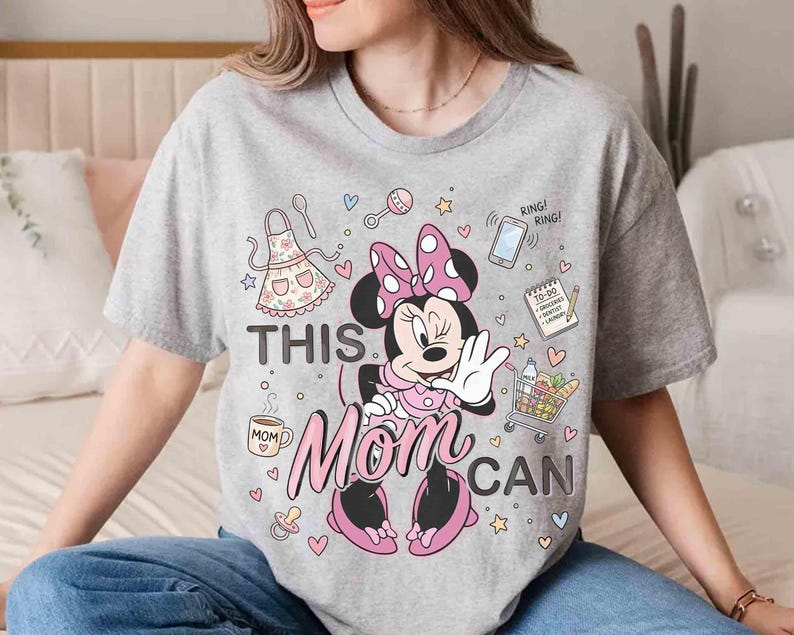 K&ouml;nnte beinhalten: Ein graues T-Shirt mit einem Minnie Mouse-Motiv und dem Text "THIS MOM CAN". Das Design umfasst eine rosa Schleife, eine Sch&uuml;rze, einen Einkaufswagen, ein Telefon und andere Symbole. Das Shirt ist ein l&auml;ssiges Kleidungsst&uuml;ck f&uuml;r den Alltag.