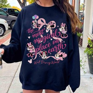 K&ouml;nnte beinhalten: Marineblaues Sweatshirt mit Minnie Mouse-Illustrationen und dem rosa Text "the most magical place on earth". Das Design umfasst eine gro&szlig;e rosa Schleife und die Worte "Walt Disney World".