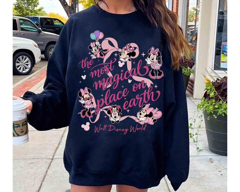 K&ouml;nnte beinhalten: Marineblaues Sweatshirt mit Minnie Mouse-Illustrationen und dem rosa Text "the most magical place on earth". Das Design umfasst eine gro&szlig;e rosa Schleife und die Worte "Walt Disney World".