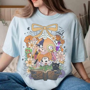 K&ouml;nnte beinhalten: Hellblaues T-Shirt mit einem Halloween-Motiv. Das Design zeigt Zeichentrickfiguren, Blumen, eine schwarze Katze und ein Zauberbuch. Die Grafik ist von einer Schleife umrahmt.