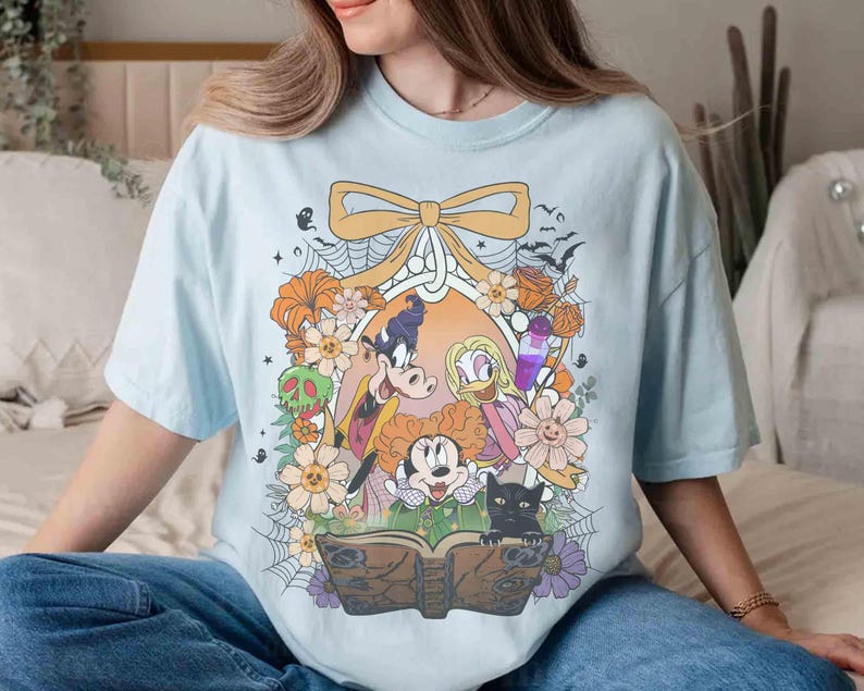 K&ouml;nnte beinhalten: Hellblaues T-Shirt mit einem Halloween-Motiv. Das Design zeigt Zeichentrickfiguren, Blumen, eine schwarze Katze und ein Zauberbuch. Die Grafik ist von einer Schleife umrahmt.