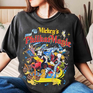 K&ouml;nnte beinhalten: Dunkelgraues T-Shirt mit einer farbenfrohen Grafik von Mickey's PhilharMagic. Das Design zeigt Mickey Mouse beim Dirigieren, Aladdin und Jasmin auf einem fliegenden Teppich und andere Disney-Charaktere. Der Text "Mickey's PhilharMagic" wird in einer lebendigen, mehrfarbigen Schrift angezeigt.