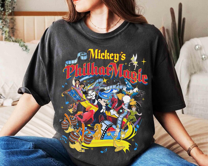 K&ouml;nnte beinhalten: Dunkelgraues T-Shirt mit einer farbenfrohen Grafik von Mickey's PhilharMagic. Das Design zeigt Mickey Mouse beim Dirigieren, Aladdin und Jasmin auf einem fliegenden Teppich und andere Disney-Charaktere. Der Text "Mickey's PhilharMagic" wird in einer lebendigen, mehrfarbigen Schrift angezeigt.