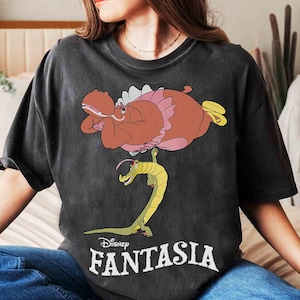 May include: Dark gray t-shirt featuring a cartoon illustration from Disney's Fantasia. The design includes a brown hippo in a pink tutu and a yellow alligator. The word "Fantasia" is printed in white below the image.