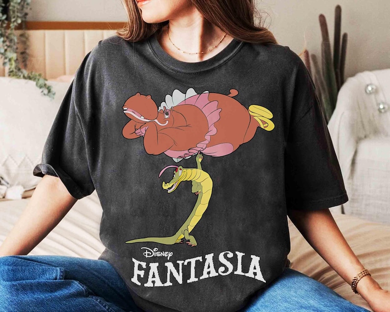 May include: Dark gray t-shirt featuring a cartoon illustration from Disney's Fantasia. The design includes a brown hippo in a pink tutu and a yellow alligator. The word "Fantasia" is printed in white below the image.